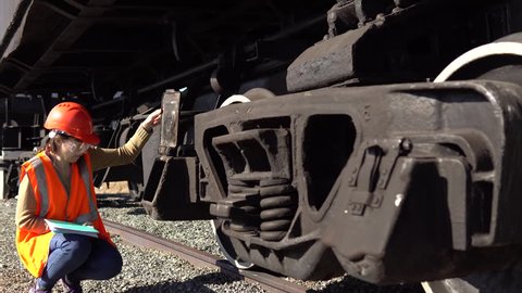 A young brunette woman railway worker in an orange vest checks the hitch unit of the freight wagon, raises the lid of the axle-box with a plain bearing and compares it with the scheme.