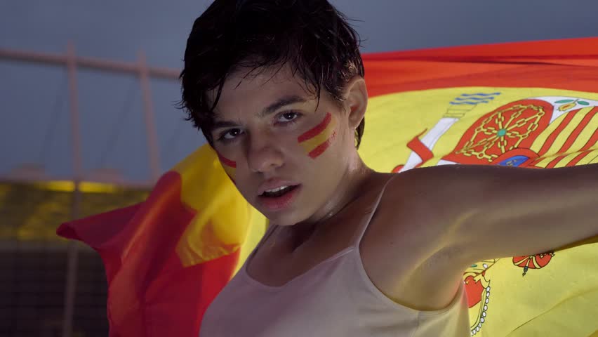 Portrait of a young brunette girl soccer fan in Spain, in the rain, looking at the camera, serious face, holding a flag behind her beck, evening stadium in the background 50 fps