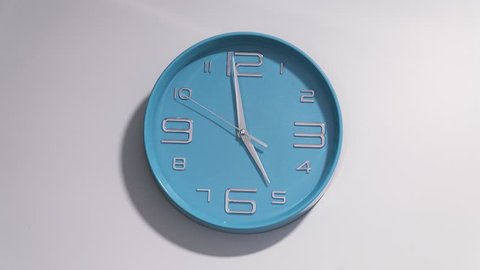 Timelapse of a blue clock on a white wall. The clock starts ticking at 5 and ends at 12.