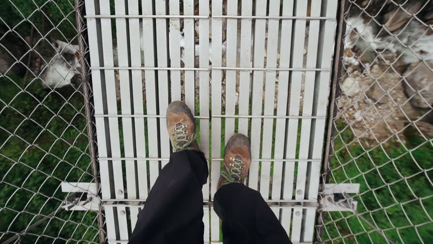 POV tourist walking through the footbridge. Traveler crossing the river on the bridge, going to the village in mountains. Slow-motion.