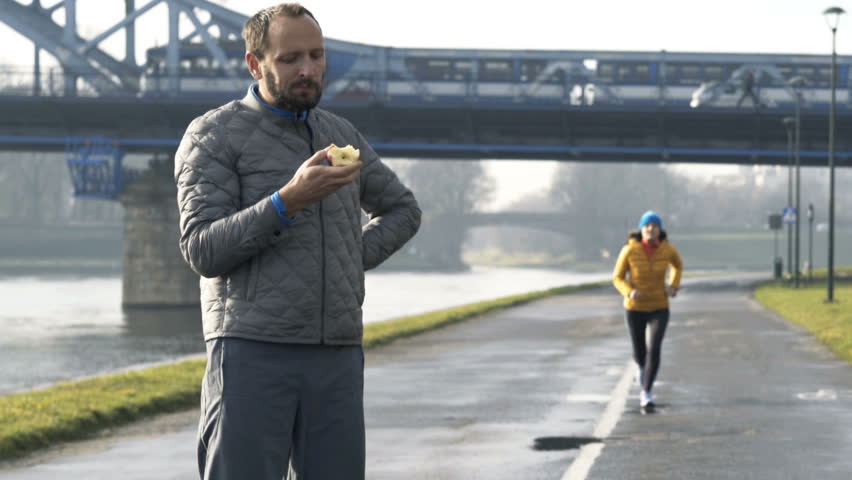 Young, sportive man eating apple and woman jogging in the city
