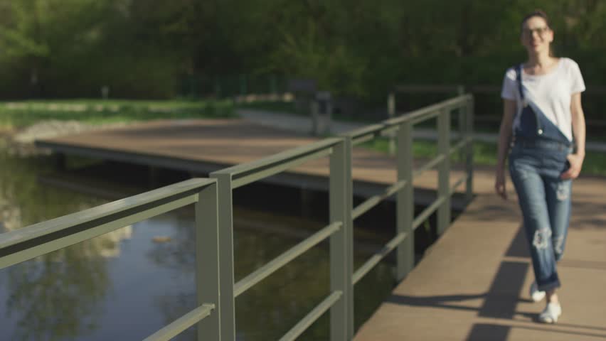 Attractive young female in eyeglasses standing in park near handrail above water and looking at camera.