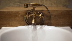 Water flows from the mouth of an old-fashioned. brass faucet. into a white. ceramic bath tub. Ultra HD 4k video