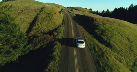 Aerial view of car driving down country road through rural rolling hills at sunset
