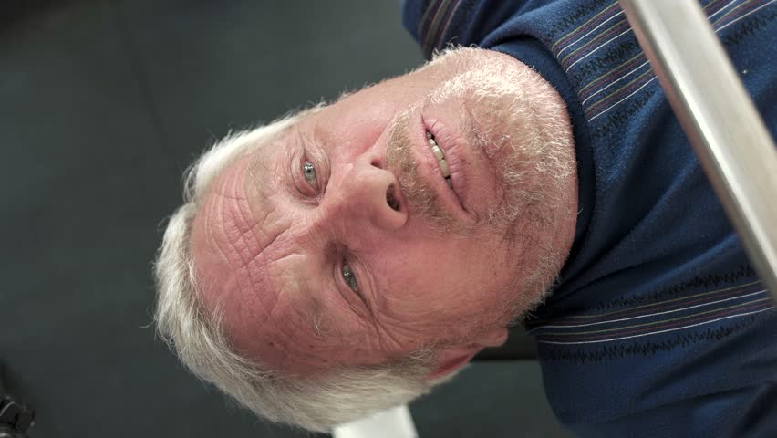 Face of elderly man lifting weights. Senior grey haired man doing heavy weight exercise for chest with barbell in gym.