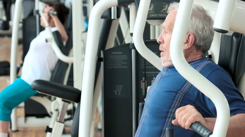 Elderly male person training at gym. Senior man exercising on machine at fitness club, side view. Active way of life.