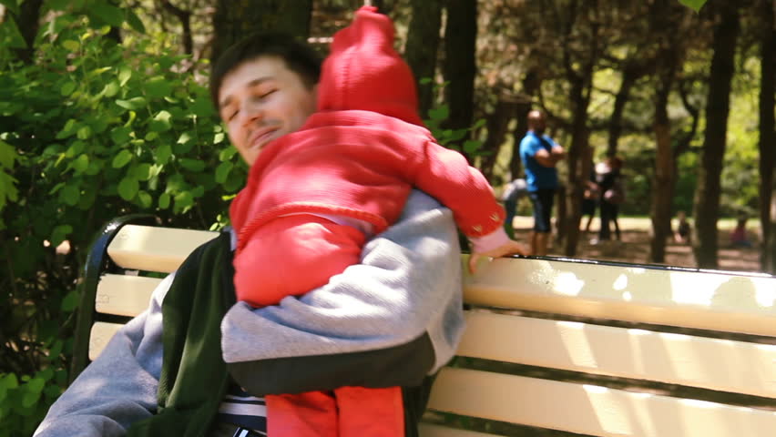 Dad and his two year old daughter. A black-haired man with a short haircut and pretty girl in a red jacket are sitting on a park bench. Dad hugs and gently cradles the baby.