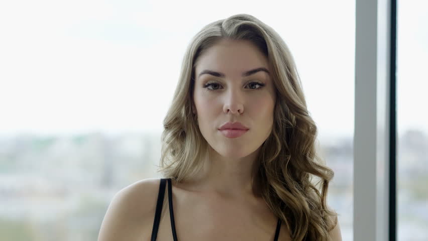 Portrait of beautiful blonde young woman in black lingerie standing at window in a luxury room. 4K