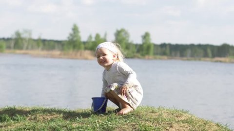 little girl with white hair sitting on the river bank, looking into the distance and playing in the sand