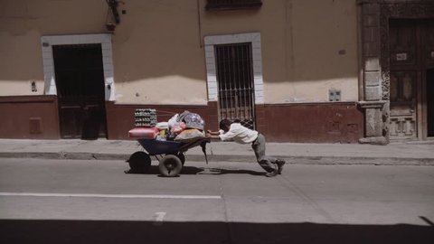 Old man pushing up the wheelbarrow loaded with groceries on the inclined street. 