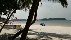 Tracking shot of tropical beach with people in sunny day calm blue water 5 Islands on background, colorful umbrellas jetski and boats in scene, tropical trees in forground raw footage
