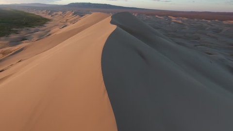 Aerial drone shot flying over the crest of a giant sand dune in gobi desert Mongolia. Sunrise time, oasis on the left and mountains in background. Low altitude flight.