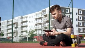 Happy teenage boy with headphones are using gadget, smiling while sitting on the playground outdoors. Young student teen with a skateboard playing on tablet pc and listening to music or watches video.