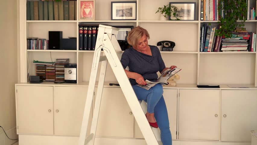 Beautiful senior woman reading newspaper on the library ladder. 
