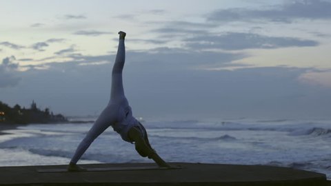 Side view of woman in white leggings and top performing standing split yoga pose on beach in morning วิดีโอสต็อก