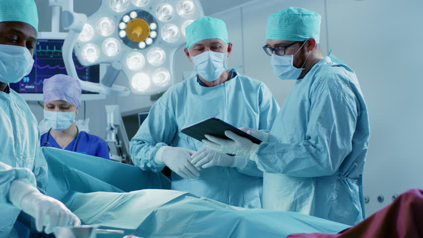 Professional Surgeons and Assistants Talk and Use Digital Tablet Computer During Surgery. They Work in the Modern Hospital Operating Room. Shot on RED EPIC-W 8K Helium Cinema Camera.