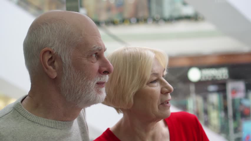 Shallow depth of field. Focus on senior man. Happy senior couple riding elevator up in modern mall. Lift going up, stores on background