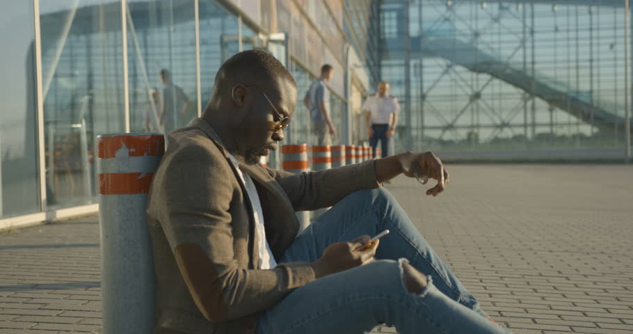 Side portrait of the confident stylish african man emotionally talking and making gestures while talking via the mobile phone, sitting on the asphalt and leaning on the road pole.