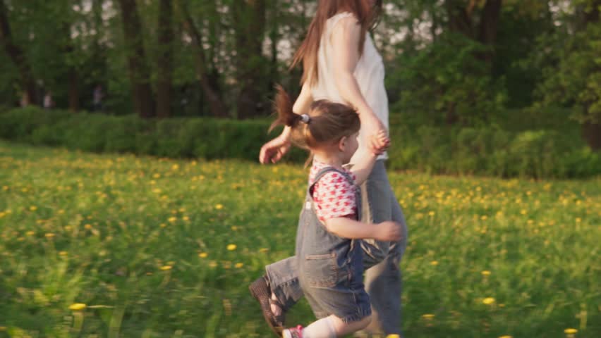 Mother and little daughter holding hands and running together on grass, slow motion, steadicam shot