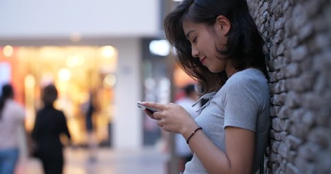 Close up Side view of one pretty young happy asian woman typing on mobile phone leaning on the wall with people walking in street at background,  Chinese woman looking at smartphone in hand,4k