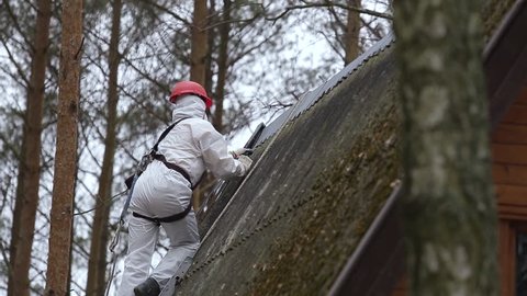 Man in a protective suit pries asbestos material off of a roof of a rural home in a forest