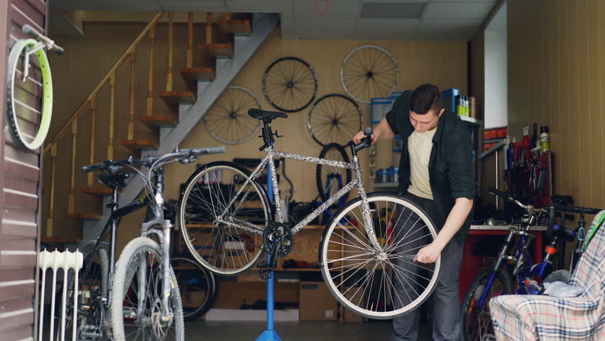 Skilled male mechanic is fixing broken bicycle wheel with wrench then turning it and checking. Many cycles, spare parts, tools and equipment are visible. | Shutterstock HD Video #1011118052
