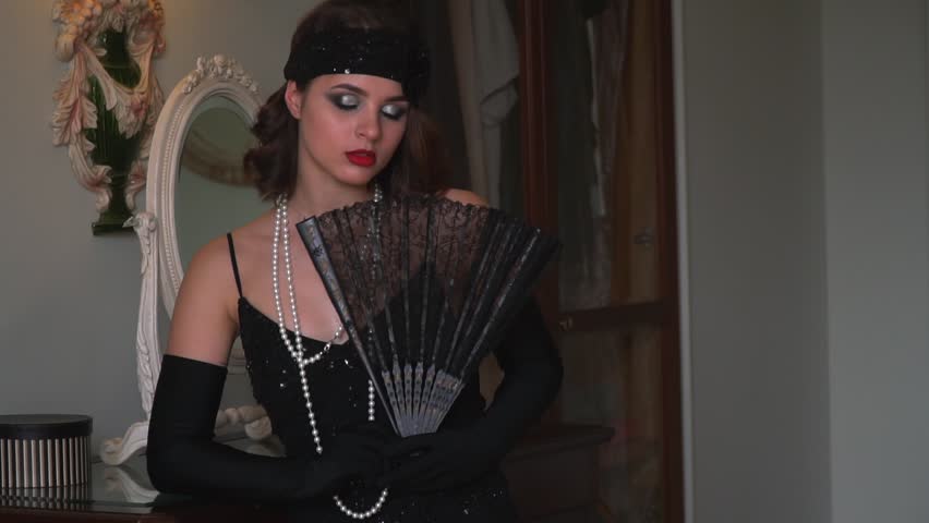 A young actress in a retro dress with a pearl necklace on her neck holds a fan in her hands
