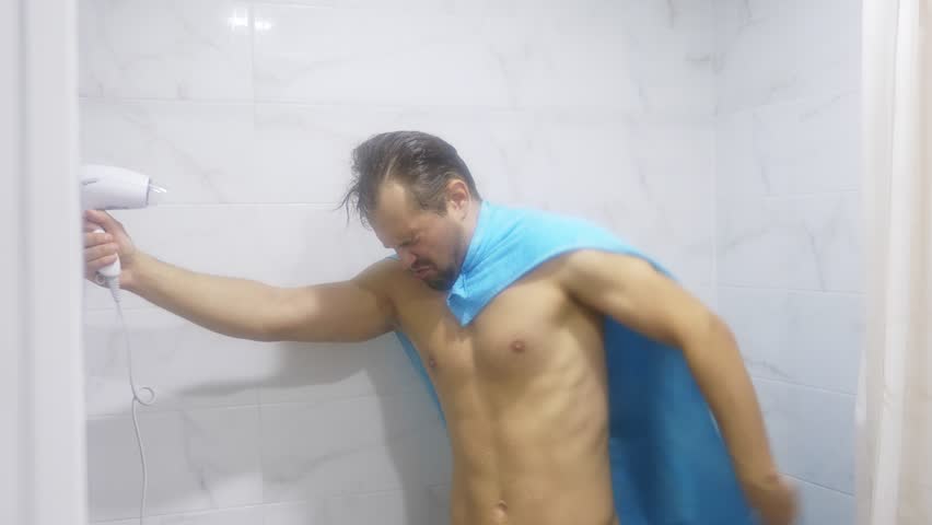 Young handsome muscular man after shower in the bathroom. He put on a towel on his shoulders, portraying a superman, and blowing himself a blow dryer. 4k | Shutterstock HD Video #1011160130