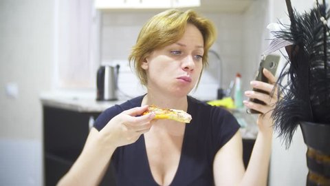 woman is eating pizza in the kitchen at home and using her smartphone, 4k, background blur