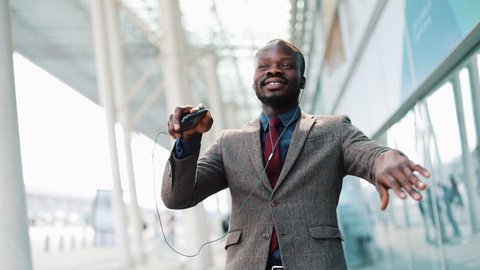 Happy african american businessman listening to music in headphones on smartphone, walking outside the office and funny dancing to the rythm. Drink coffee, funny dancing moves. Close up