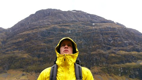 4K Man in the Scottish Highlands. Person looking up and standing on mountains. Hiker in hooded yellow jacket and backpack in a travel rural tourist destination spot