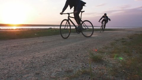 Two young men riding bicycles on the beach on the background of an orange sunsetting sky, slow motion