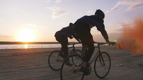 Two young men riding bicycles on the beach with orange smoke during sunset, slow motion