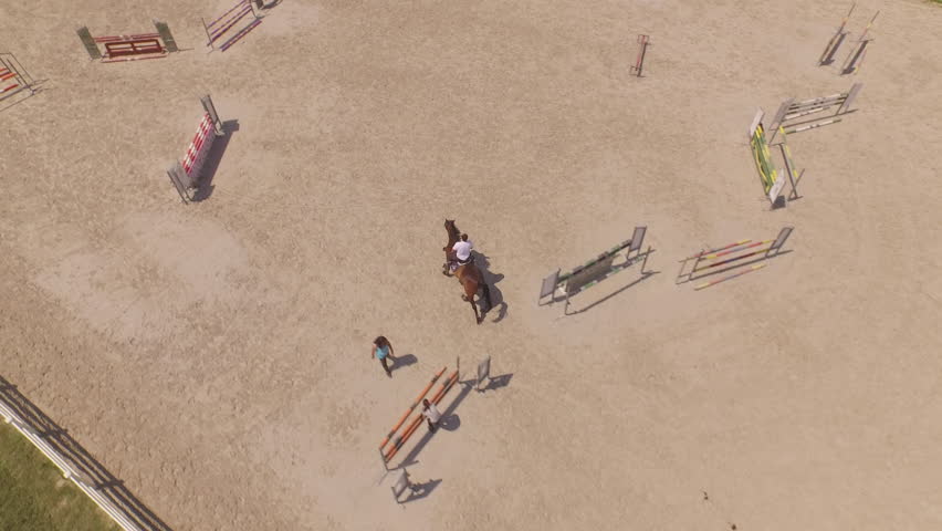 AERIAL: Flying above male sport rider riding dark bay gelding showjumping over fence obstacles in big professional sandy parkour dressage arena. Horseman competing in equestrian jumping discipline Royalty-Free Stock Footage #1011186797