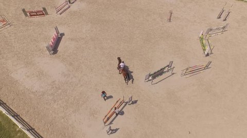 AERIAL: Flying above male sport rider riding dark bay gelding showjumping over fence obstacles in big professional sandy parkour dressage arena. Horseman competing in equestrian jumping discipline