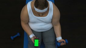 Obese man lifting dumbbell and watching fitness video smartphone, green screen