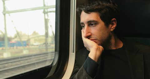 Young man scratching face while looking out window while riding and commuting by train