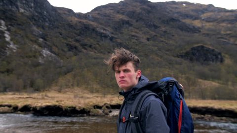 4K Young traveller hiker portrait looking at camera in front of Steall waterfall Scottish Highlands UK. A man backpacking nature path  and trekking through mountains in Scotland in slow motion 