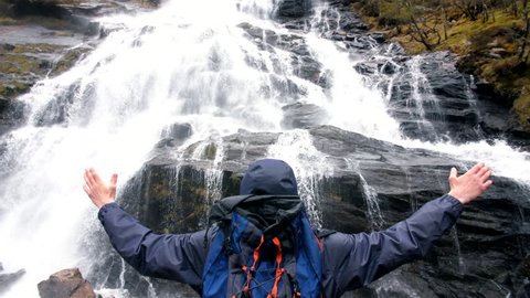 4K Young male traveller standing in front of Steall waterfall in Scottish Highlands. Slow motion man embracing nature with arms open wide in the air. Backpacker hiker in Scotland UK 
