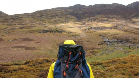 4K Hiking traveller looking over Scottish Highland landscapes. Young man with yellow raincoat and backpack on walking up mountain in Scotland UK 