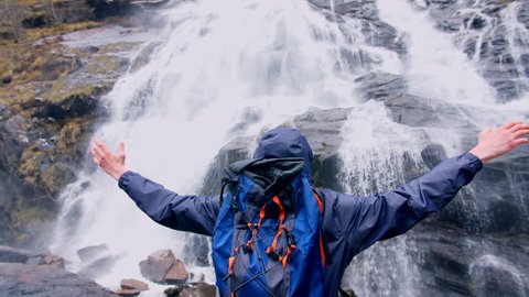 4K Young man Hiker with backpack raising arms in front of Steall waterfall in Scottish Highlands. Backpacker and traveller in Scotland UK. Mountain landscapes in slow motion. 