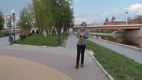 young man plays the saxophone. man on the background of the city's landscape blows into the pipe. musician goes to the camera along the embankment of the river.