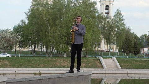 young man plays the saxophone. The musician plays on the wind instrument, walks the parapet to the camera, the camera is in motion. Man moves on the narrow road. man on the background of the city