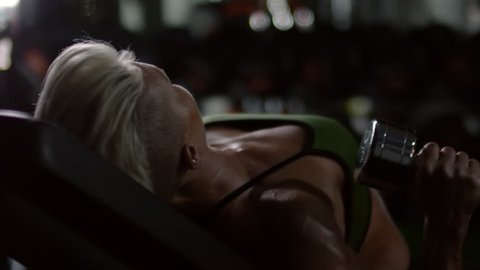 Closeup of muscular woman lying on bench and doing dumbbell press exercise in dark gym