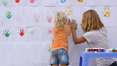 A little cute happy funny child painting color handprints on the white wall with smiling mother together and giving five by hands in print 50fps