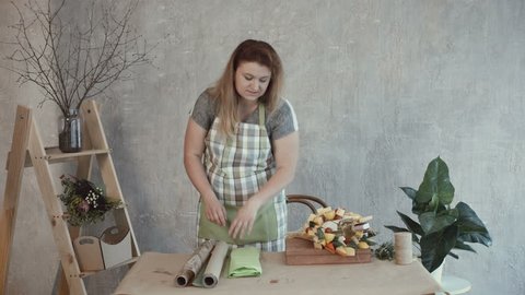 Attractive woman in apron choosing wrapping paper for edible bouquet arrangement at workplace. Creative positive redhead female making edible arrangement and choosing craft paper.
