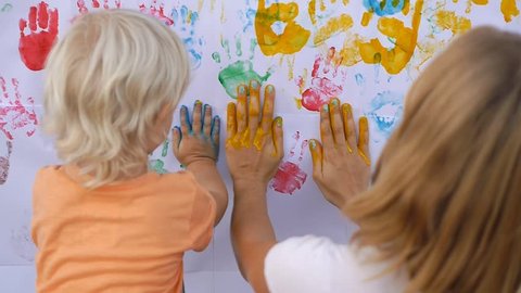 Cute little child painting color handprints on the white wall with mother together. 50fp
