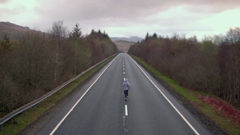 4K of a man jogging on a road in the Scottish Highlands. Tree lined country side British landscape of a runner with a cloudy sky and mountain horizon.