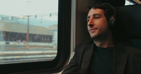 Man riding train listening to music, podcast, or audiobook while riding train  and looking out window. Young man removing headphones