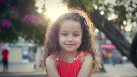 Close-up portrait of a happy cute little girl with curly hair in a red dress, sits in the park and looks at the camera on the sunset background. Stock footage.